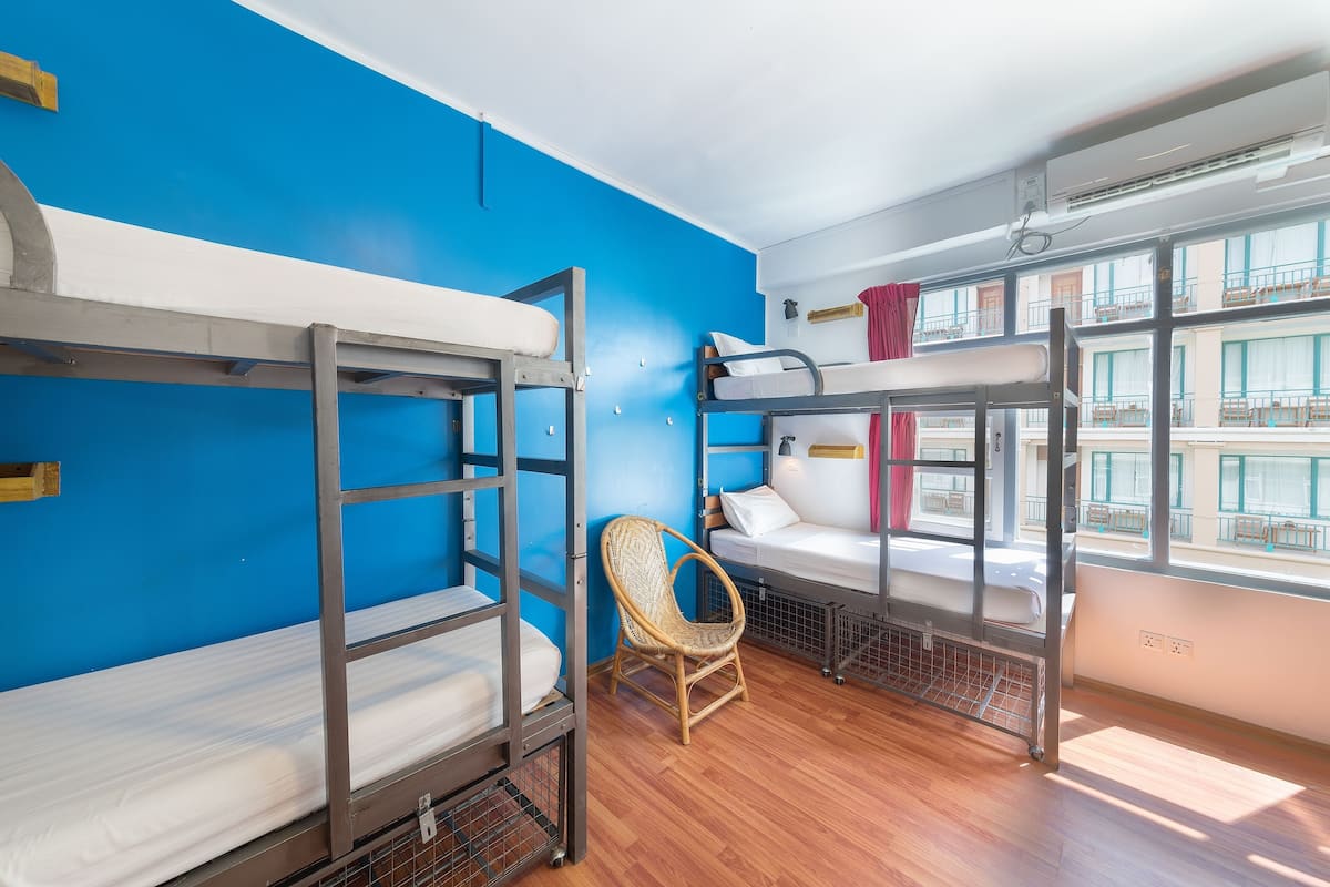 Une chambre de dortoir avec des lits superposés, une chaise en osier et une grande fenêtre donnant sur un immeuble.