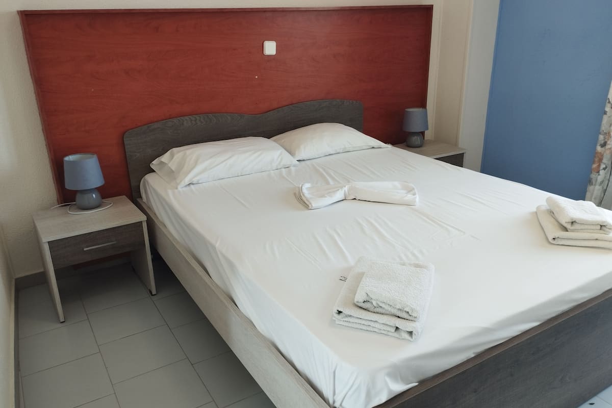 A neatly made bed with white linens and towels, a red headboard, two bedside lamps, and a small bedside table with a lamp.