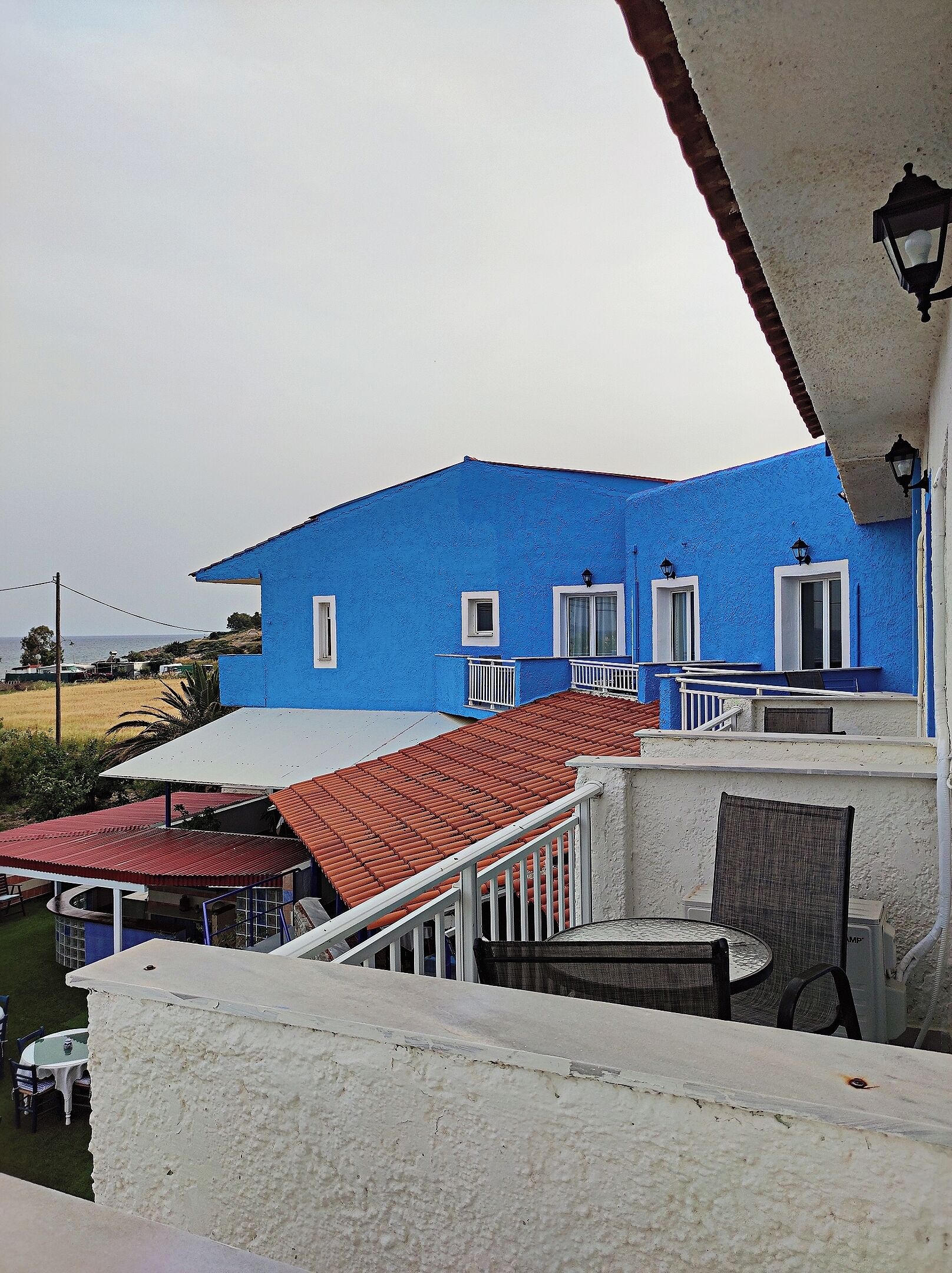 Een blauw gebouw met een rode pannendak, witte relingen en een balkon met een tafel en stoelen.