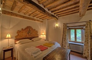 A bedroom with a wooden ceiling, a bed with white linens, a bench, a nightstand with a lamp, and a window with curtains. - Villa Cretole (Monterchi)