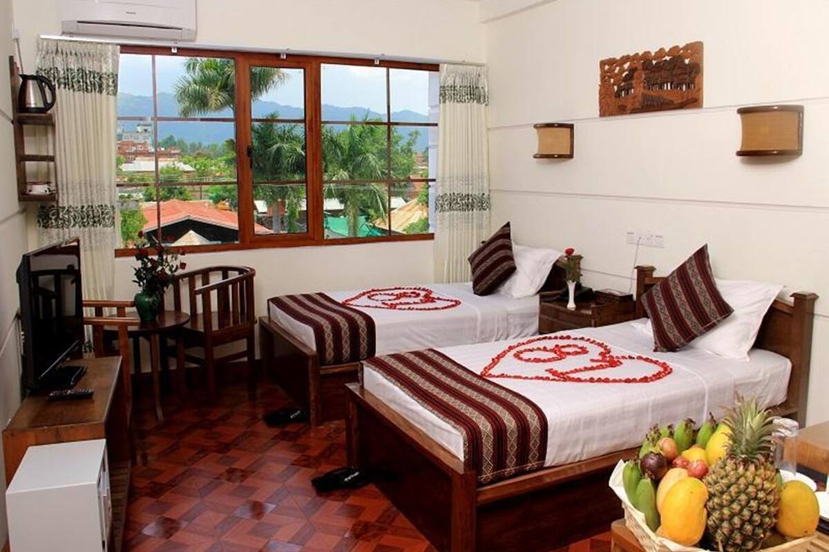 Une chambre d’hôtel avec deux lits, un bureau, une chaise, une petite table avec des fruits, et une vue sur des palmiers et des montagnes.