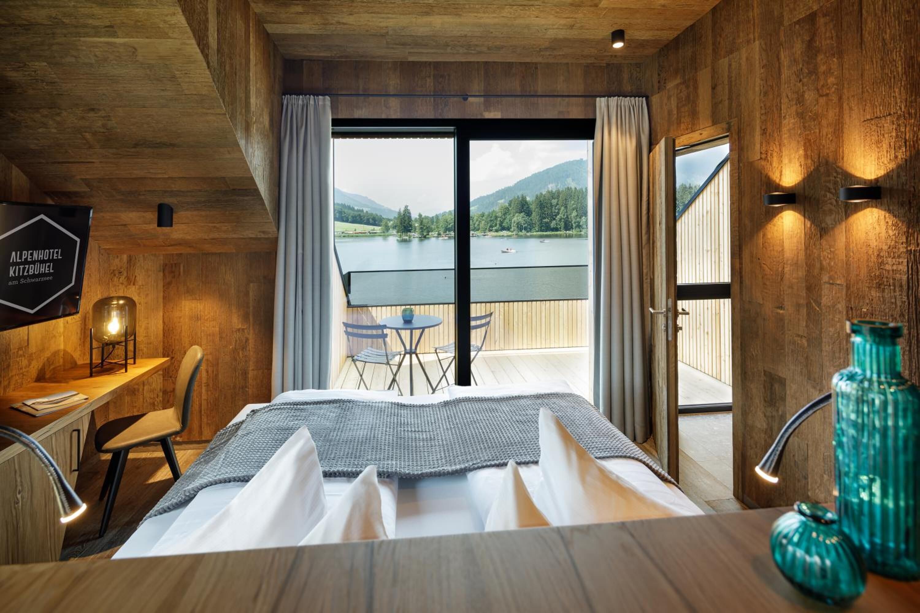 Habitación de hotel con una cama grande, un escritorio con lámpara, vista a un lago y montañas a través de las puertas del balcón, y paredes de madera.