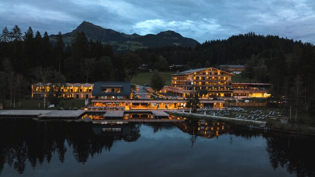A large, multi-story hotel with a reflection in the water, surrounded by trees and mountains. - Alpenhotel Kitzbühel (Kitzbuhel)