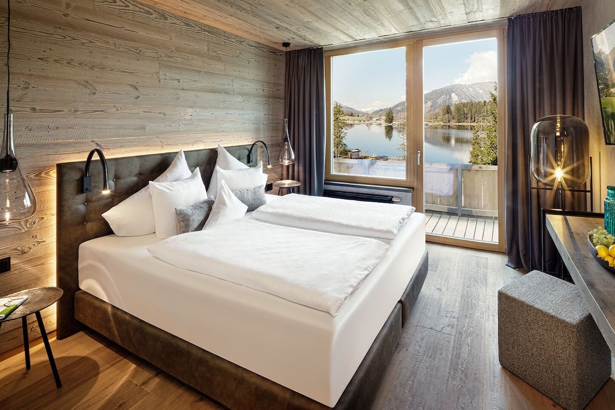 Une chambre d’hôtel moderne dotée d’un grand lit, d’une tête de lit en bois, d’une vue sur un lac et d’un coin repas avec une table et des chaises.