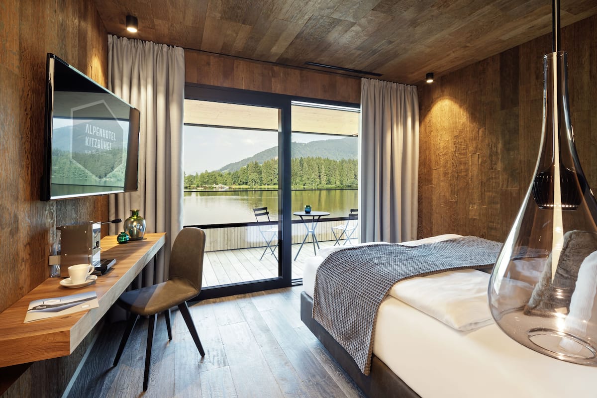 Une chambre d’hôtel moderne dotée d’un grand lit, d’un bureau avec une machine à café, d’une télévision et offrant une vue sur un lac et des montagnes.