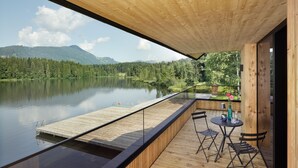 A wooden deck with a glass barrier overlooking a lake, a dock, and a table with chairs. - Alpenhotel Kitzbühel (Kitzbuhel)
