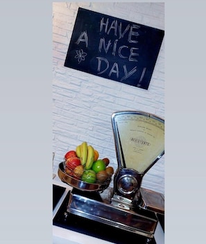 A kitchen counter with a fruit bowl containing bananas, apples, and limes, a scale, and a blackboard with 'HAVE A NICE DAY!' written on it.