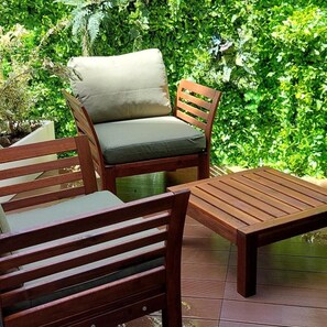 Un espace de détente extérieur en bois, avec un fauteuil rembourré et une table, le tout entouré d’une végétation luxuriante.