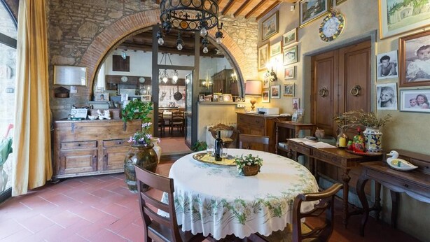 Un comedor rústico con una mesa redonda, sillas y una araña. Se observan armarios de madera y una pared de piedra al fondo.