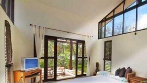 Ein Schlafzimmer mit einem großen Bett, einem Fernseher auf einem Holzbalken und Blick auf einen Garten durch eine Glastür.