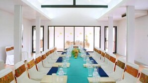 Ein Konferenzraum mit einem langen Tisch, der für eine Besprechung gedeckt ist, weißen Stühlen mit gemusterten Bändern und großen Fenstern, die für natürliches Licht sorgen.