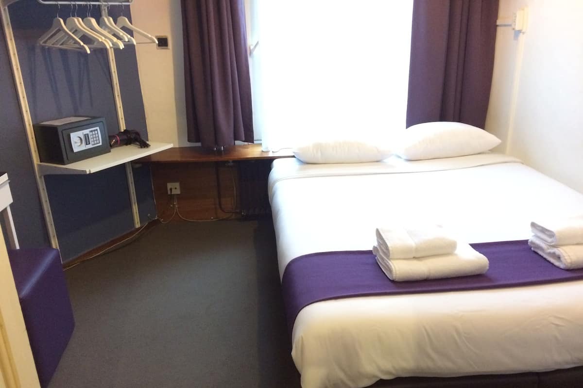 A hotel room with a bed, a desk with a radio, a wardrobe, and a window with curtains.