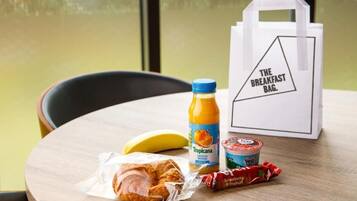 A breakfast table with a croissant, banana, Tropicana orange juice, Yeo yogurt, and a snack pack. A white bag labeled "The Breakfast Bag" is also visible.
