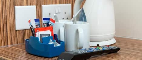 Un control remoto, una taza blanca, un portavasos azul con revistas y una tetera blanca sobre una mesa de madera.