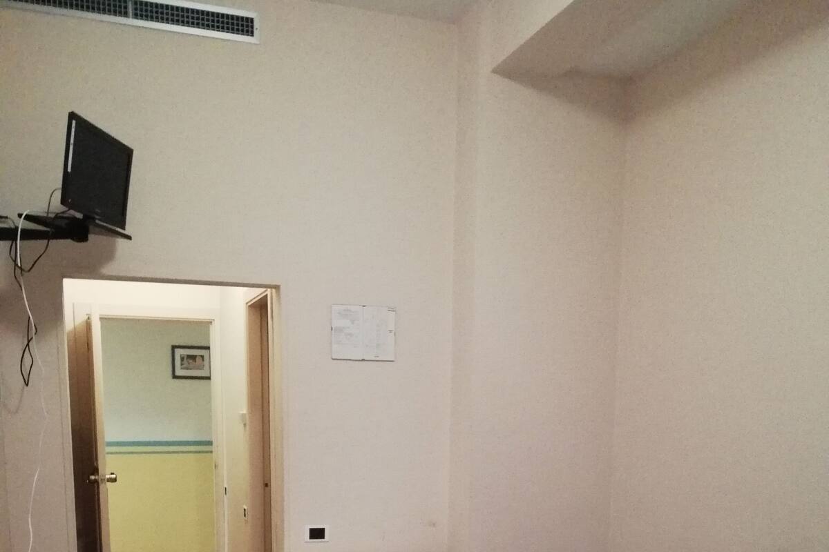 A room with a bed, a desk, a chair, a TV mounted on the wall, and a door leading to another room.