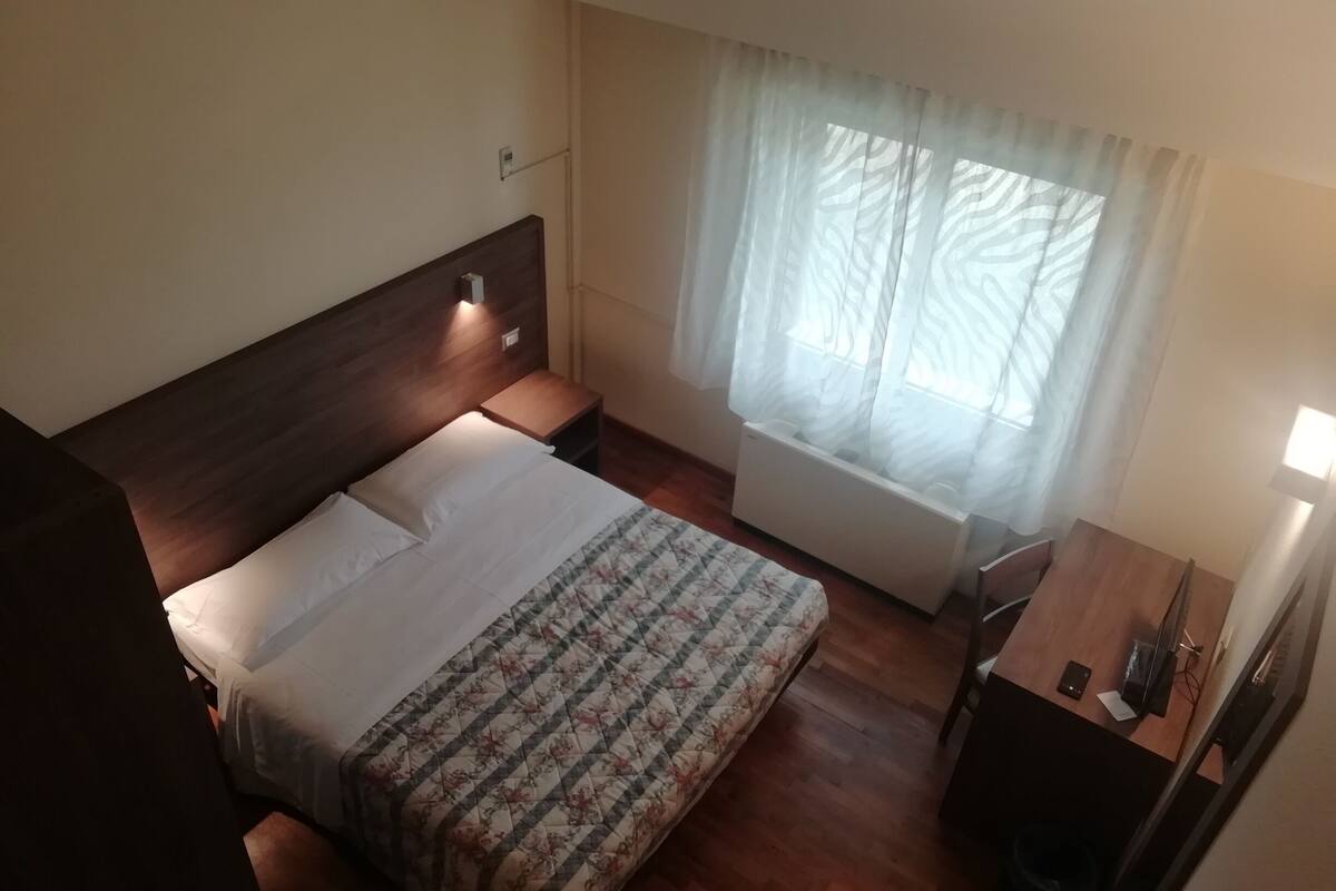 A hotel room with a bed, bedside table, lamp, and a window with sheer curtains.