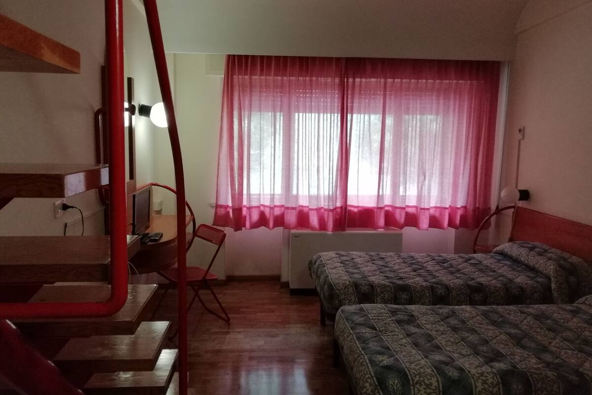 A room with two beds, a desk, a chair, a red pipe, and a window with pink curtains.