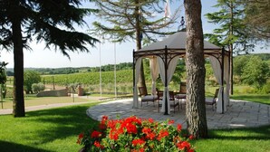 A gazebo with a table and chairs, surrounded by a garden with red flowers, set in a landscaped area with trees and a vineyard in the background. - I Grandi Di Toscana (Civitella in Val di Chiana)