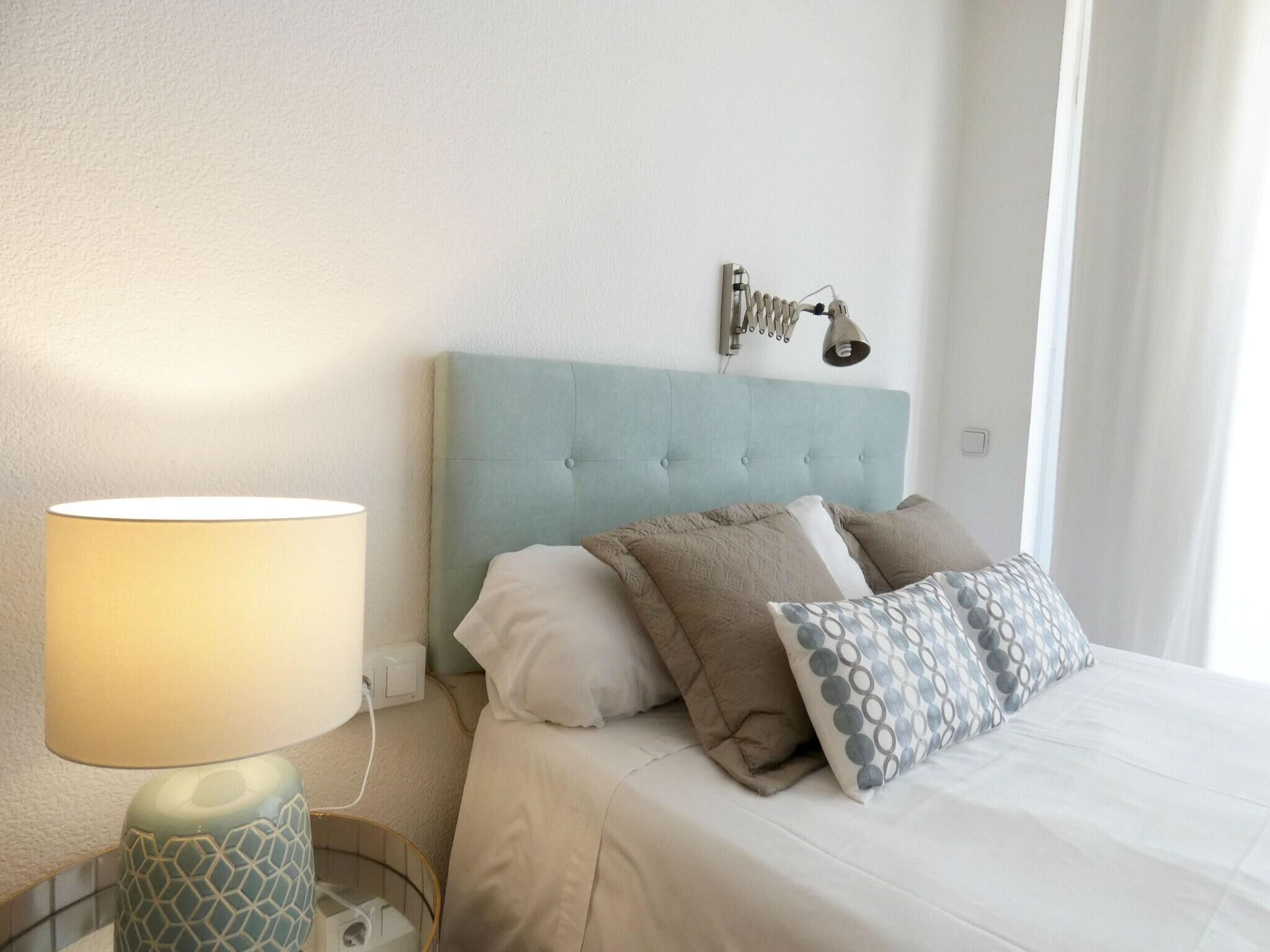 Una cama bien tendida con cabecera azul claro, una lámpara de noche y un aplique de pared.