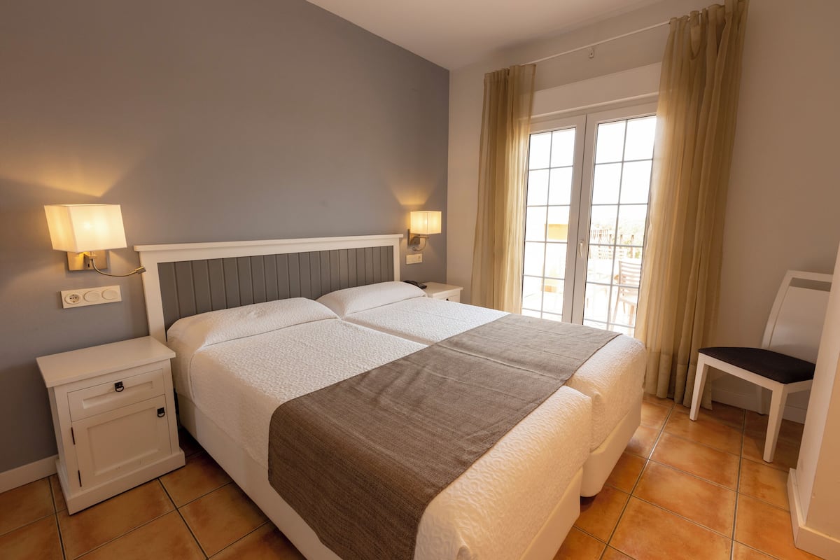 Quarto de hotel com uma cama grande, duas mesas de cabeceira, uma cadeira e uma janela com cortinas.