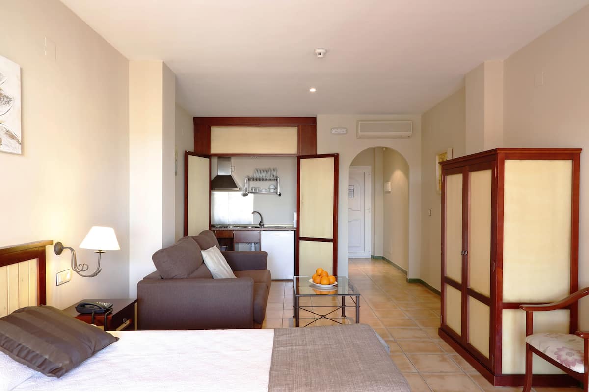 Quarto de hotel com cama, um sofá, uma mesa pequena com laranjas, uma cadeira, mesa de cabeceira com abajur e vista para a cozinha.
