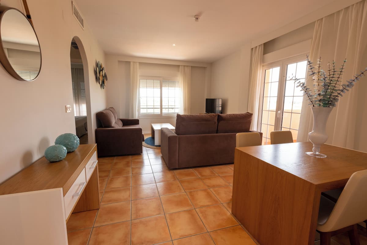 Uma sala ampla com sofá, mesa de centro e mesa de jantar. Há uma TV montada na parede e uma janela com cortinas.