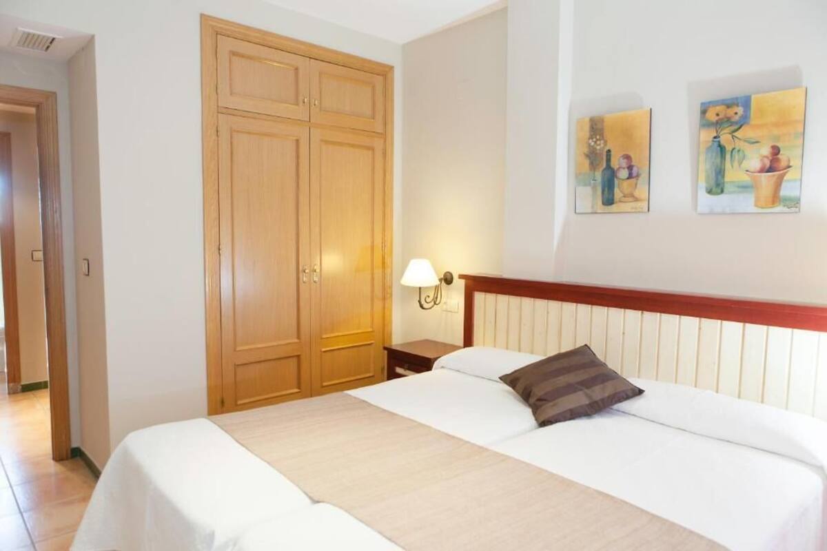 Quarto de hotel com um guarda-roupa de madeira grande, uma cama com roupa de cama branca, uma mesa de cabeceira com abajur e duas pinturas penduradas na parede.