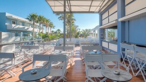 Une terrasse extérieure avec des chaises et des tables blanches, un espace salon abrité et une vue sur les palmiers et les bâtiments.