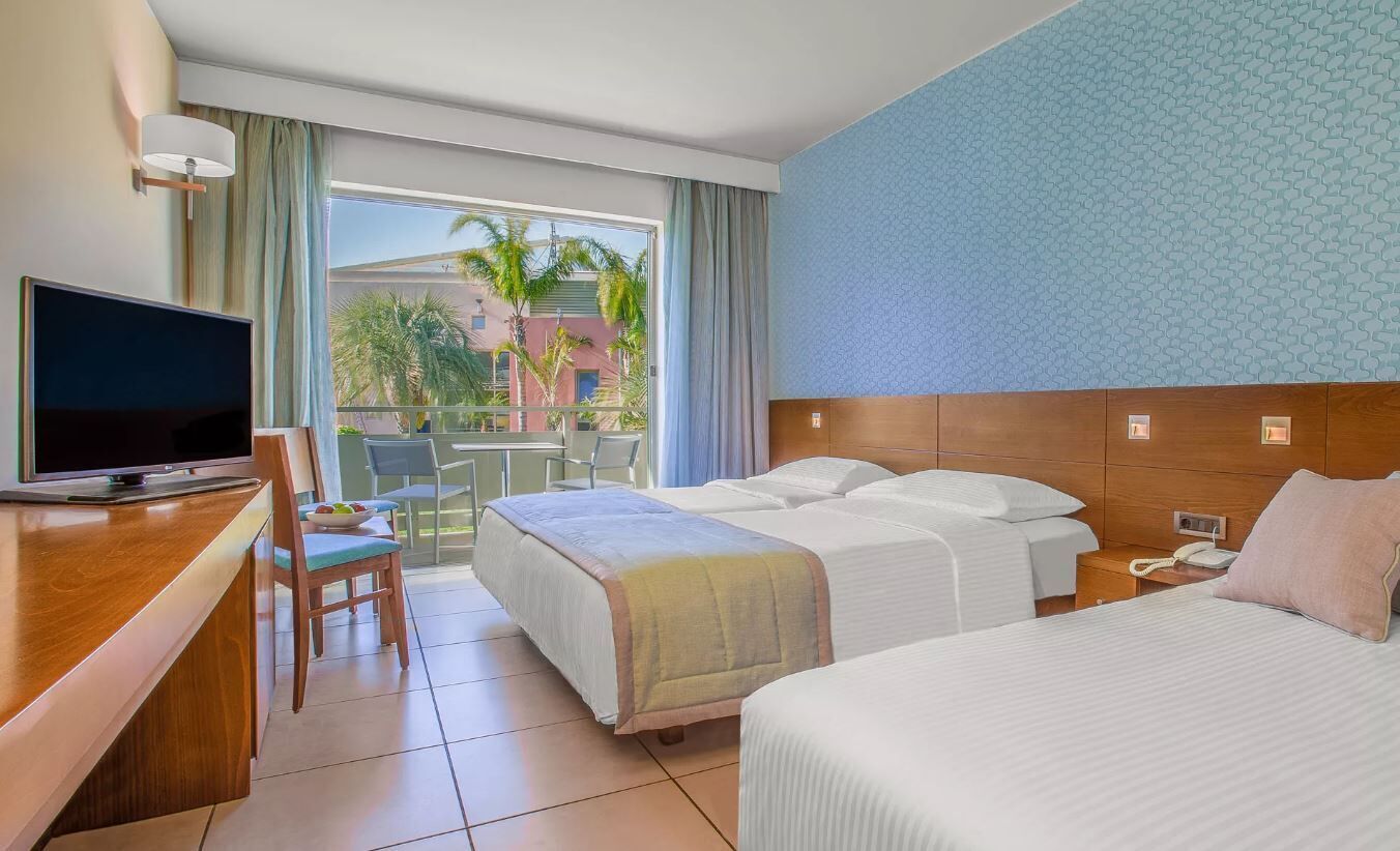 A hotel room with two beds, a TV, a desk, and a balcony with a view of palm trees.