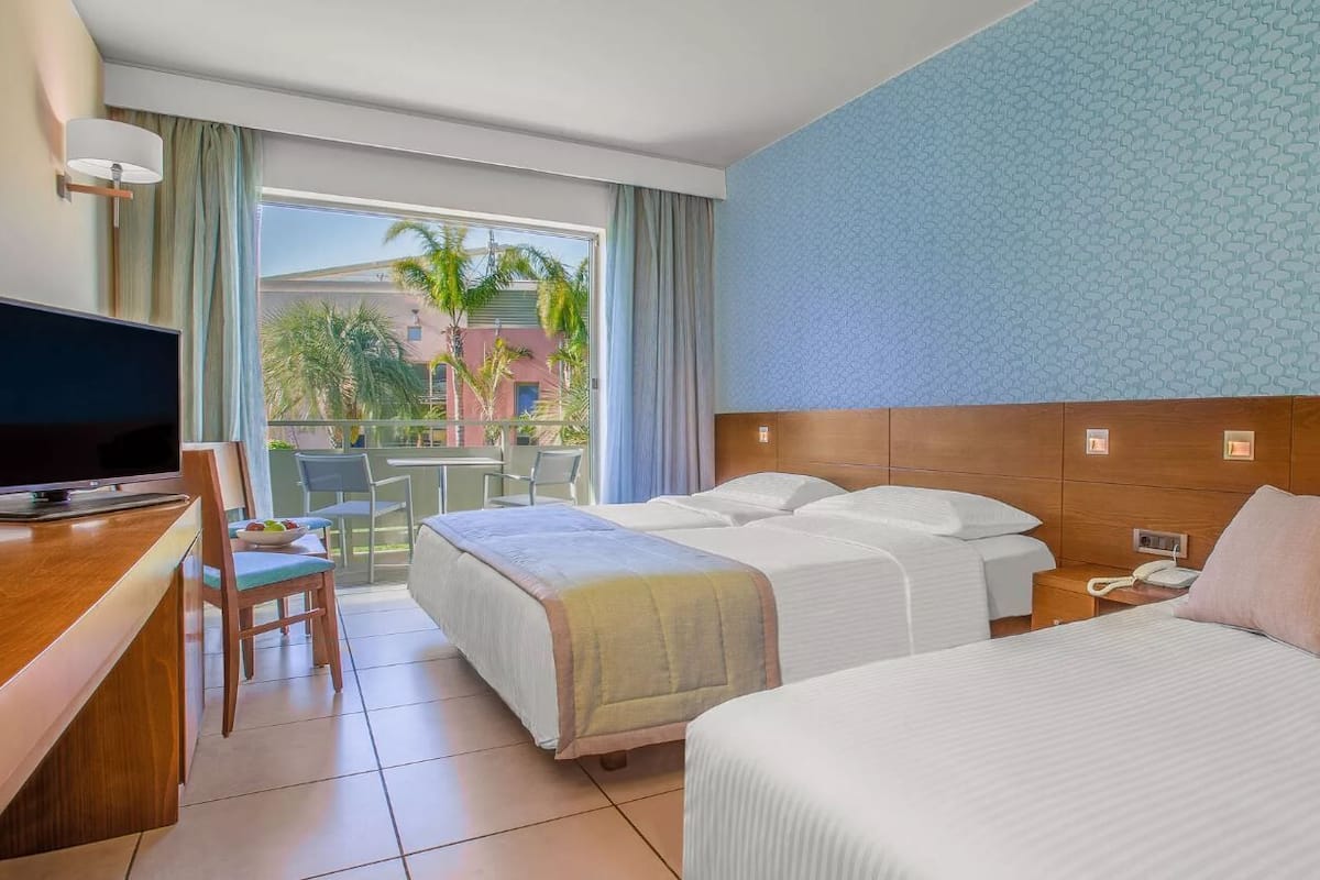 A hotel room with two beds, a TV, a desk, and a balcony with a view of palm trees.