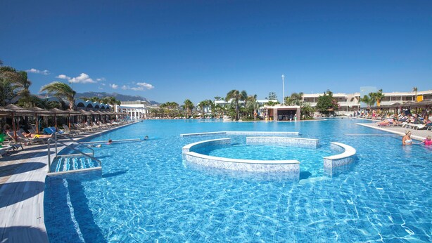 A large swimming pool with multiple lanes, surrounded by sun loungers and umbrellas, with buildings in the background.