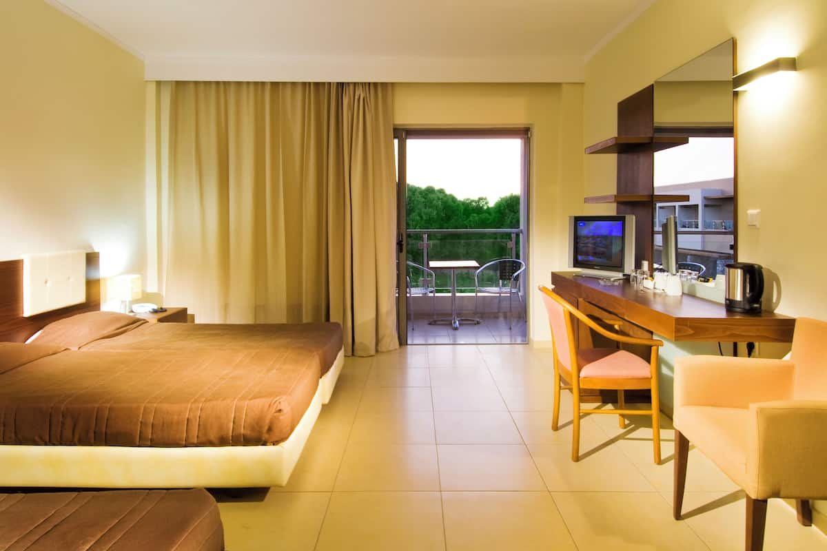 A hotel room with a bed, a desk with a chair, a television, and a balcony with a view of greenery.