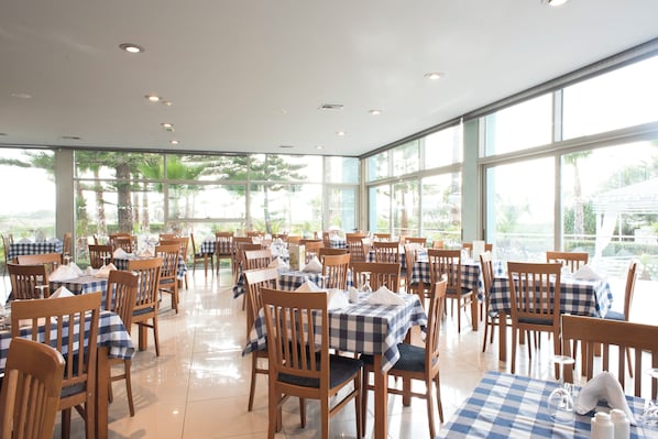 A spacious dining area with multiple tables and chairs, large windows offering a view of palm trees and greenery.