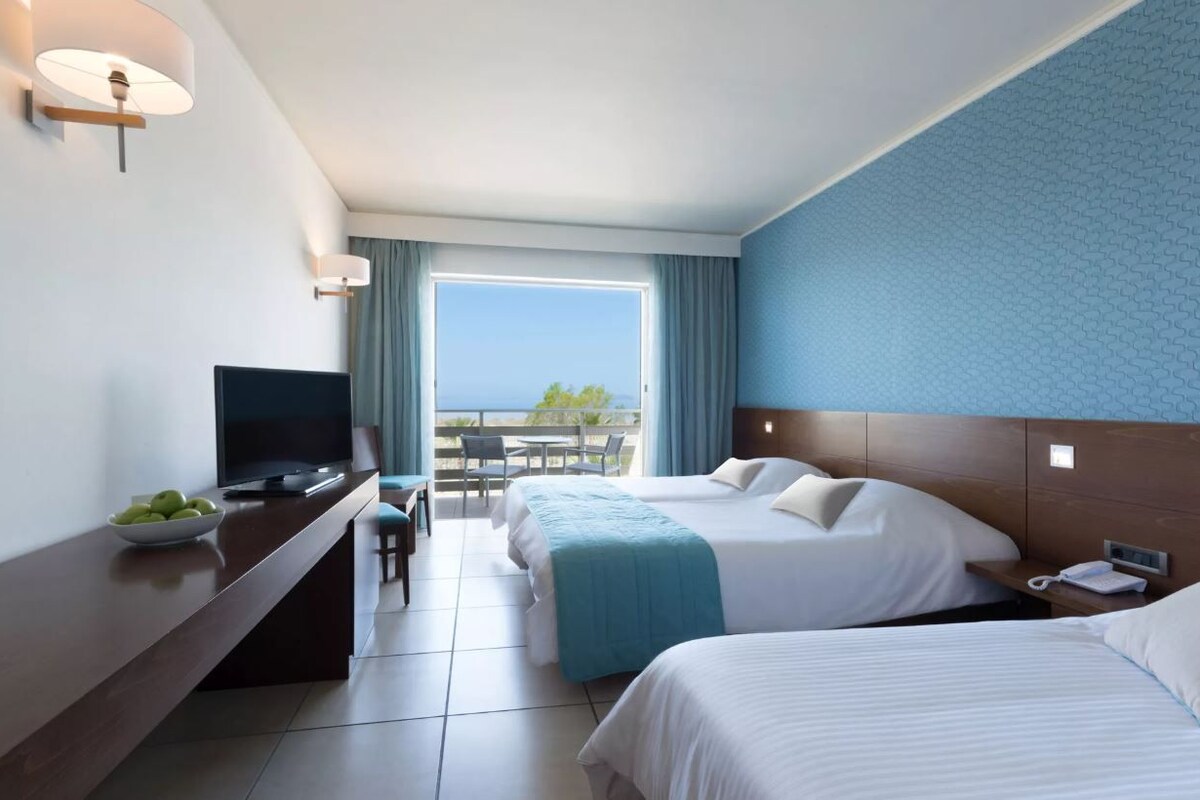 Une chambre d’hôtel avec deux lits, une télévision, un bureau avec un panier de fruits, un balcon avec vue et un téléphone sur la table de chevet.