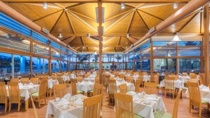 A spacious restaurant with multiple tables and chairs, large windows, and a wooden ceiling structure.