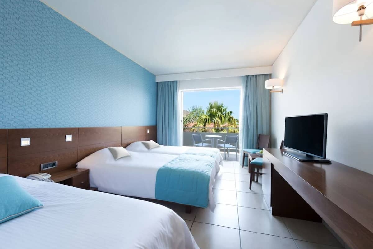 A hotel room with two beds, a TV, a desk, and a balcony with outdoor seating.