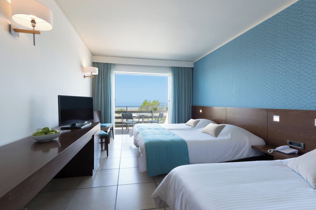 A hotel room with two beds, a TV, a desk with a bowl of fruit, a balcony with a view, and a phone on the nightstand.