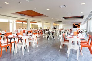 A spacious dining area with multiple tables and chairs, large windows, and a digital display on the wall. - Blue Lagoon Resort (Kos)