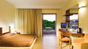 A hotel room with a bed, a desk with a chair, a television, and a balcony with a view of greenery.