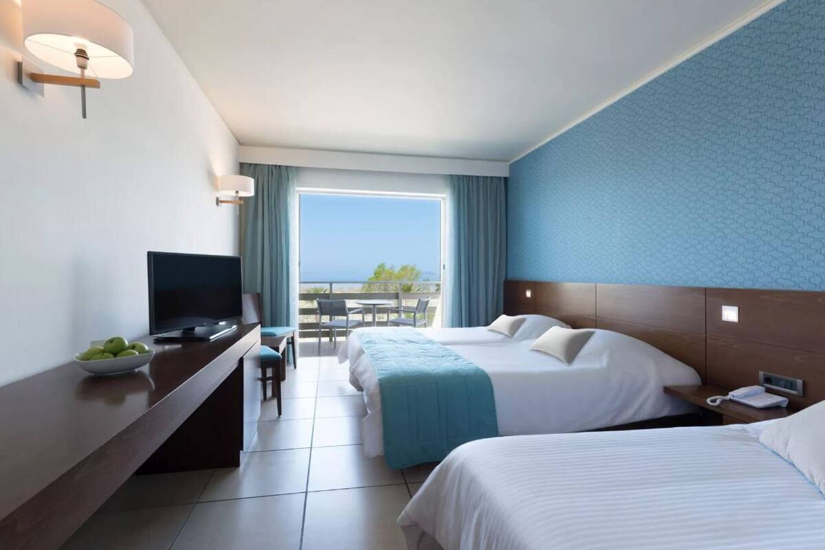 A hotel room with two beds, a TV, a desk with a bowl of fruit, a balcony with a view, and a phone on the nightstand.
