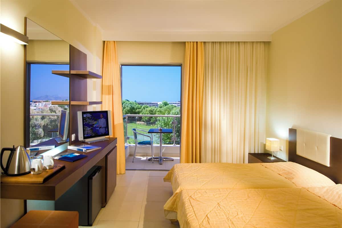 A hotel room with a bed, a desk with a computer, a chair, a balcony with a view, and a window with curtains.