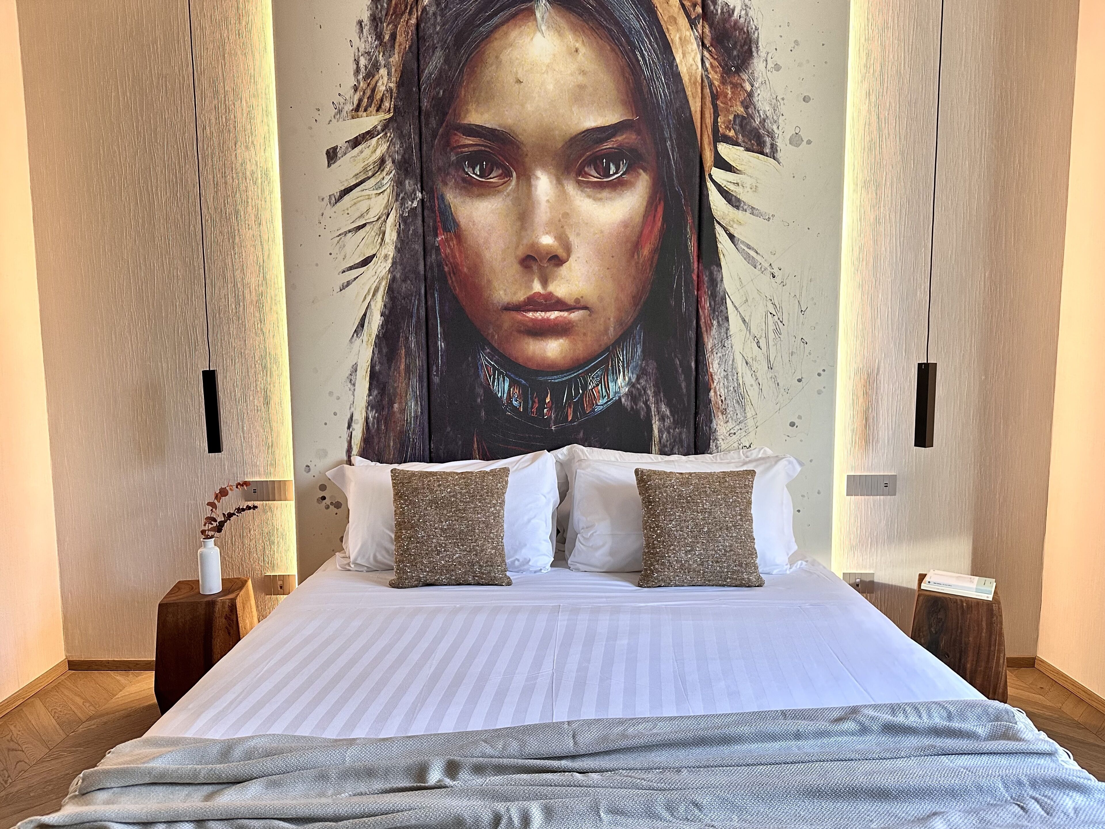 A hotel room with a large bed, two decorative pillows, a bedside table with a lamp, and a wall mural.