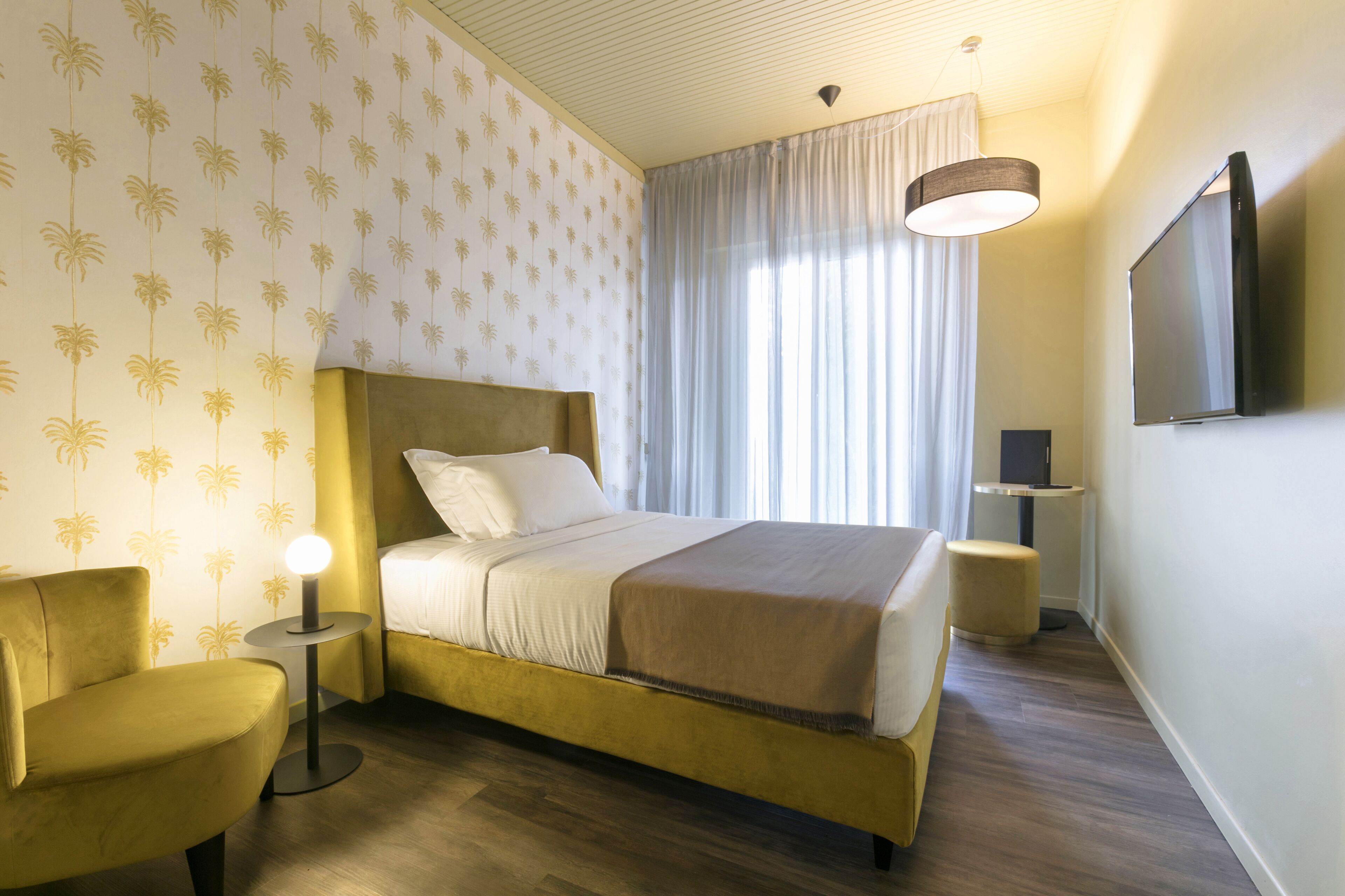 Una camera d'albergo con un letto, un comodino con una lampada, una sedia, una televisione e una finestra con tende.