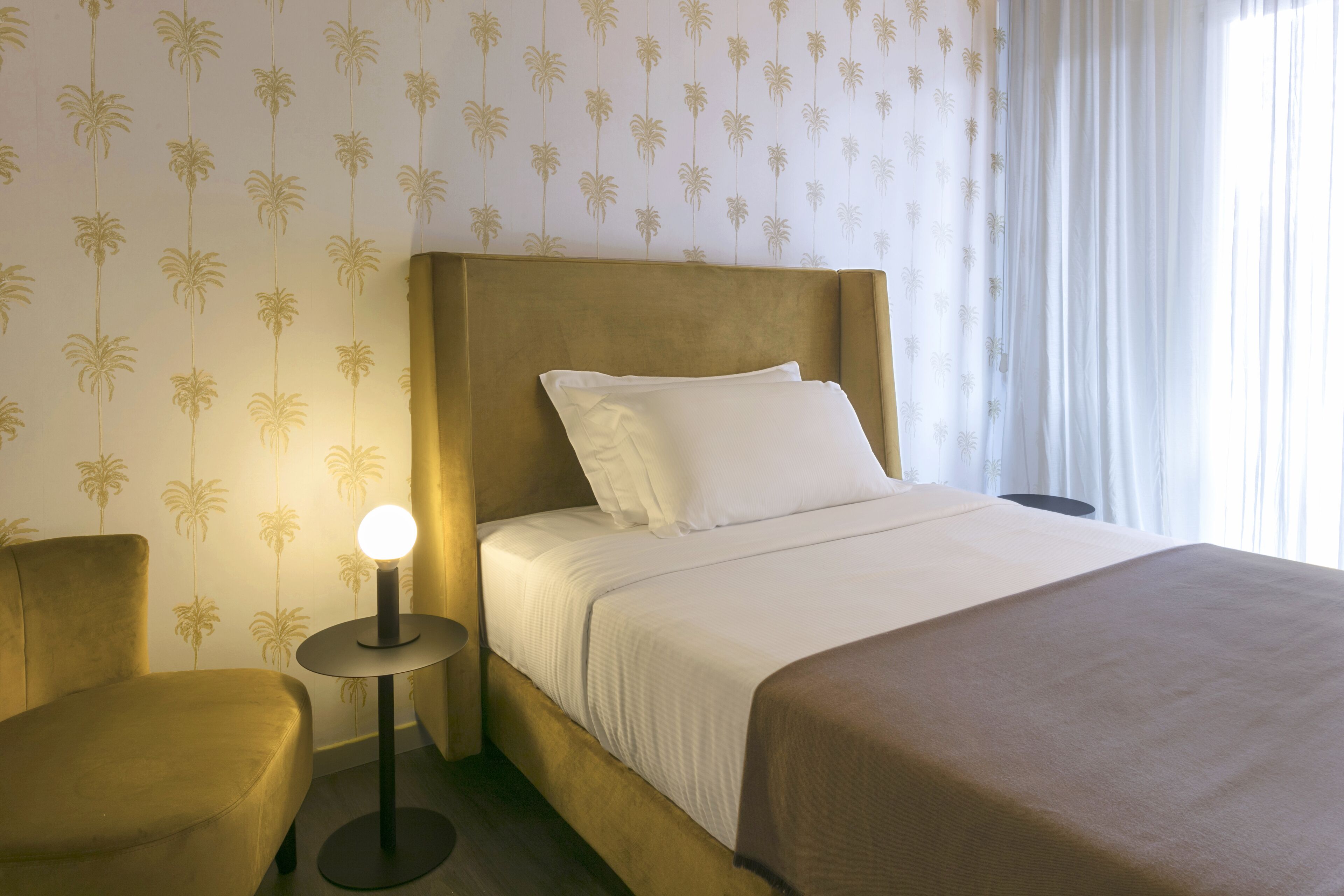 A hotel room with a bed, a bedside table with a lamp, a chair, and palm tree wallpaper.