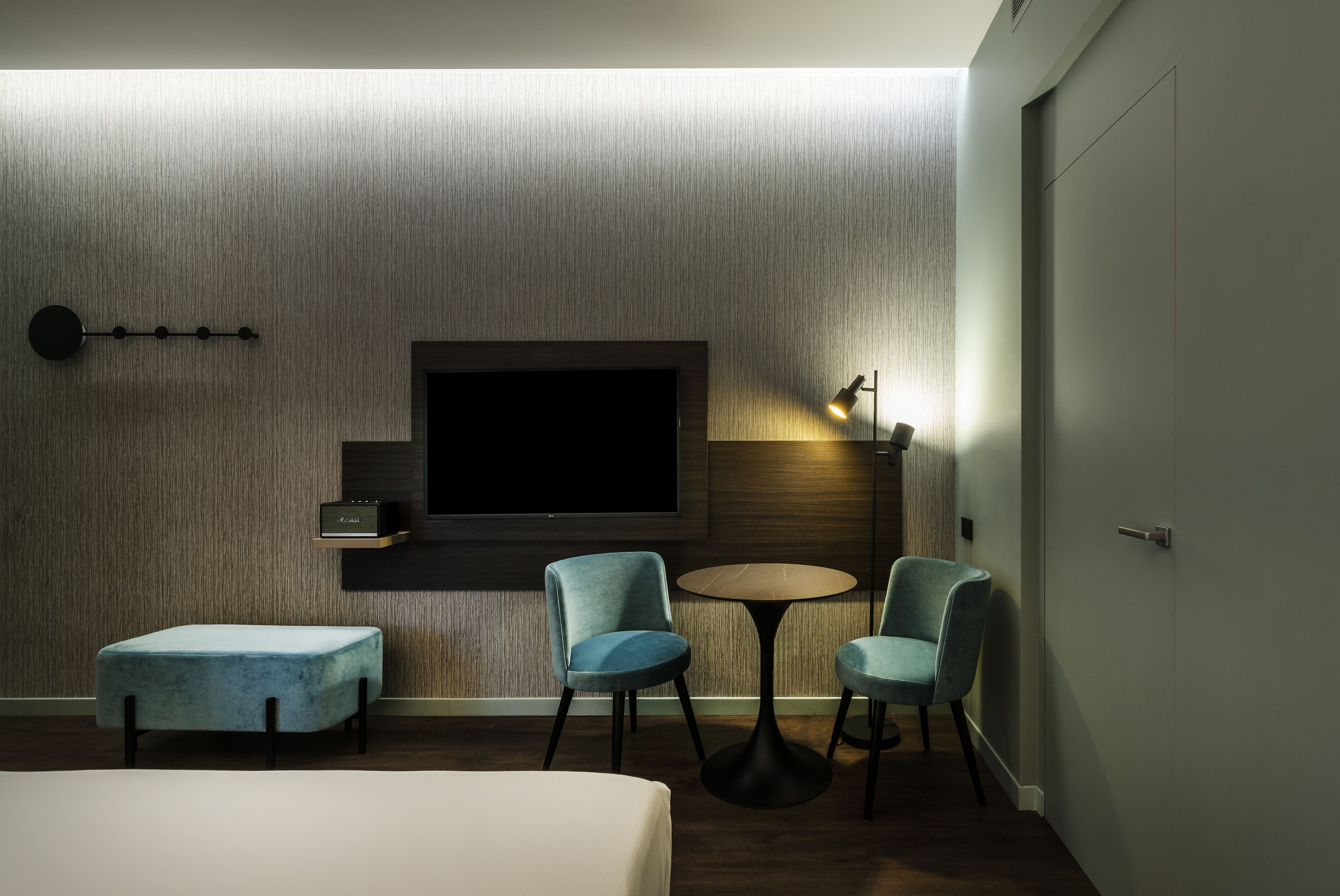 A modern hotel room with a large flat-screen TV mounted on the wall, a small table with two chairs, a cushioned bench, and a door on the right.