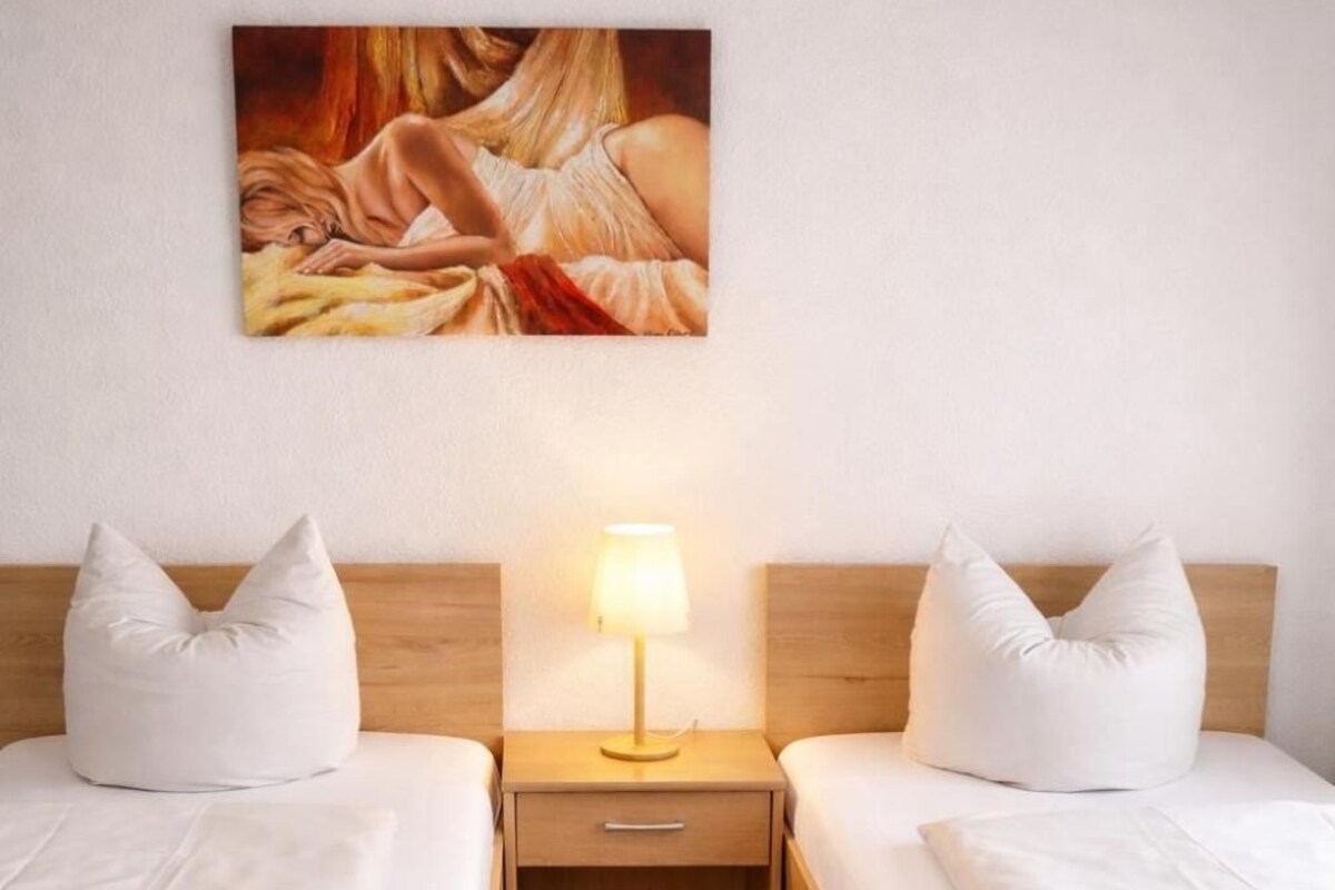 Habitación de hotel con dos camas individuales, una mesita de noche con lámpara y un cuadro en la pared.