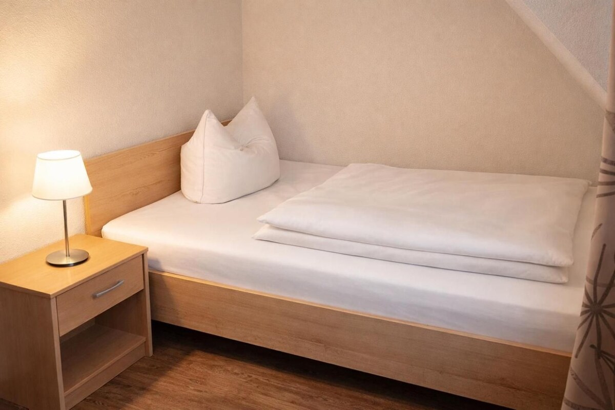 Una habitación pequeña con una cama individual, una mesita de noche, una lámpara y una cortina.
