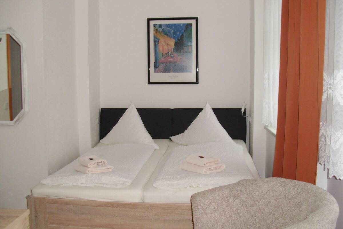 A double bed with white linens and pillows, a framed picture on the wall, and a window with curtains.