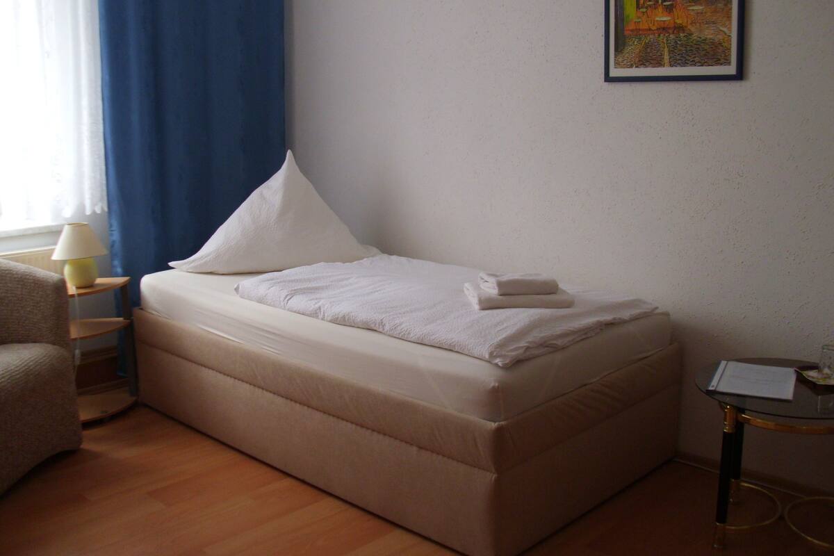A single bed with a white mattress and pillows, a bedside table with a lamp, a chair, and a framed picture on the wall.