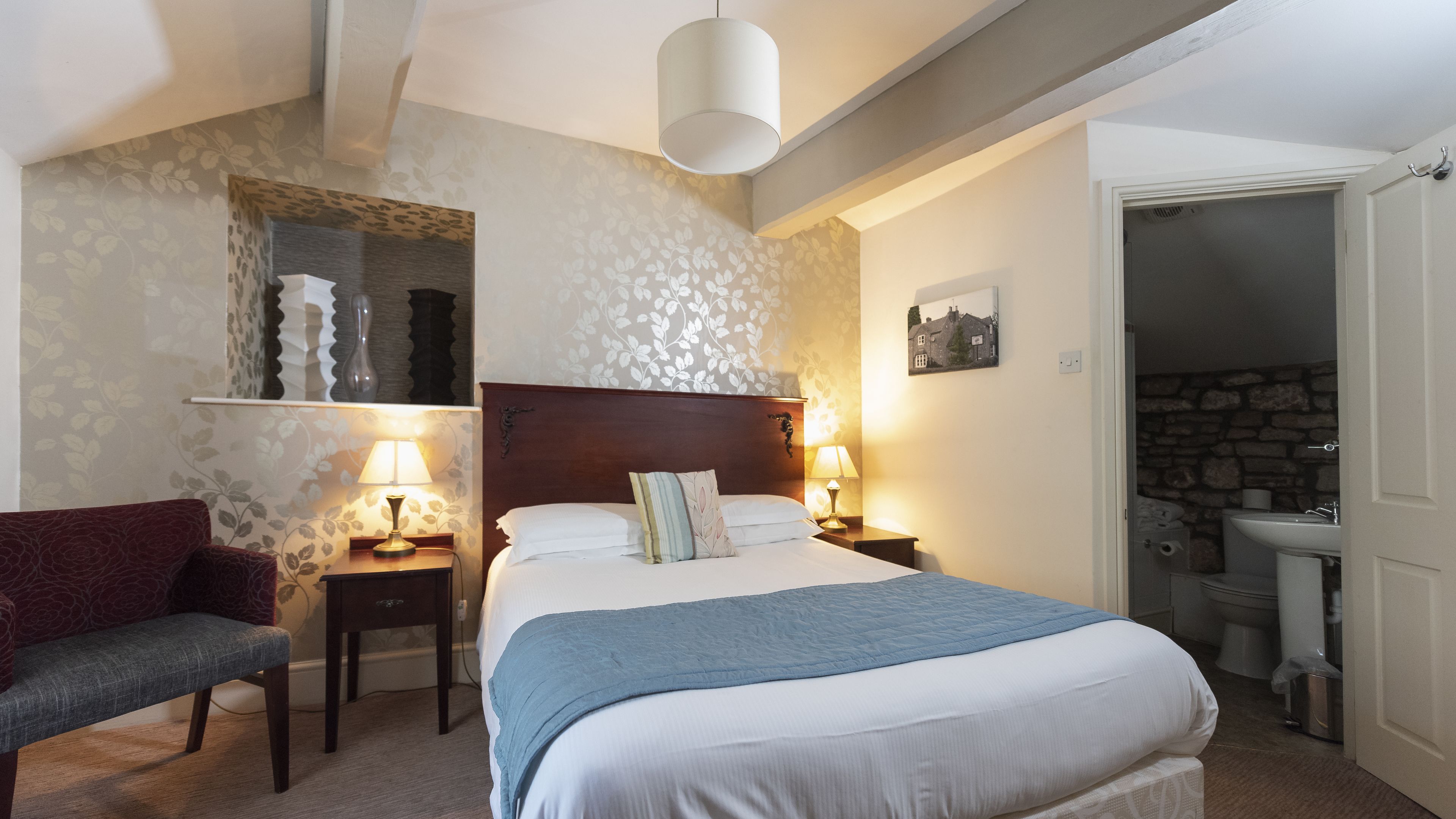 Une chambre à coucher avec un lit, deux tables de chevet avec des lampes, une chaise, un tableau accroché au mur et une salle de bain visible à travers une porte ouverte.